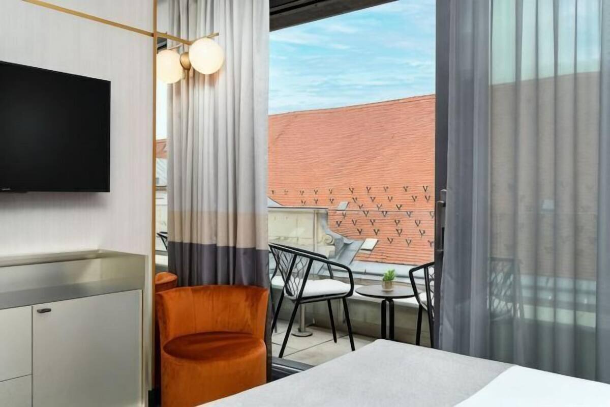 A modern hotel room with a TV, a dining area with chairs, and a view of a brick building.