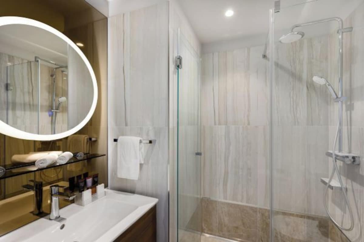 Un baño moderno con ducha de vidrio, espejo redondo y lavamanos blanco con artículos de aseo.