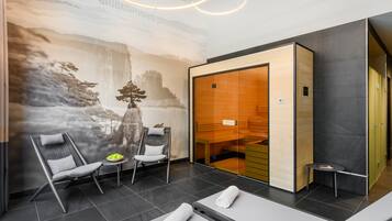 A modern spa room with a large mural, two massage tables, a seating area with chairs, and a small table with a bowl of fruit.