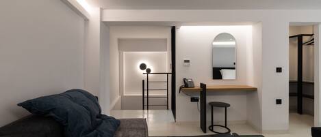 A modern hotel room with a glass table, a sofa with a dark blanket, a small stool, a desk with a phone, and a mirror.