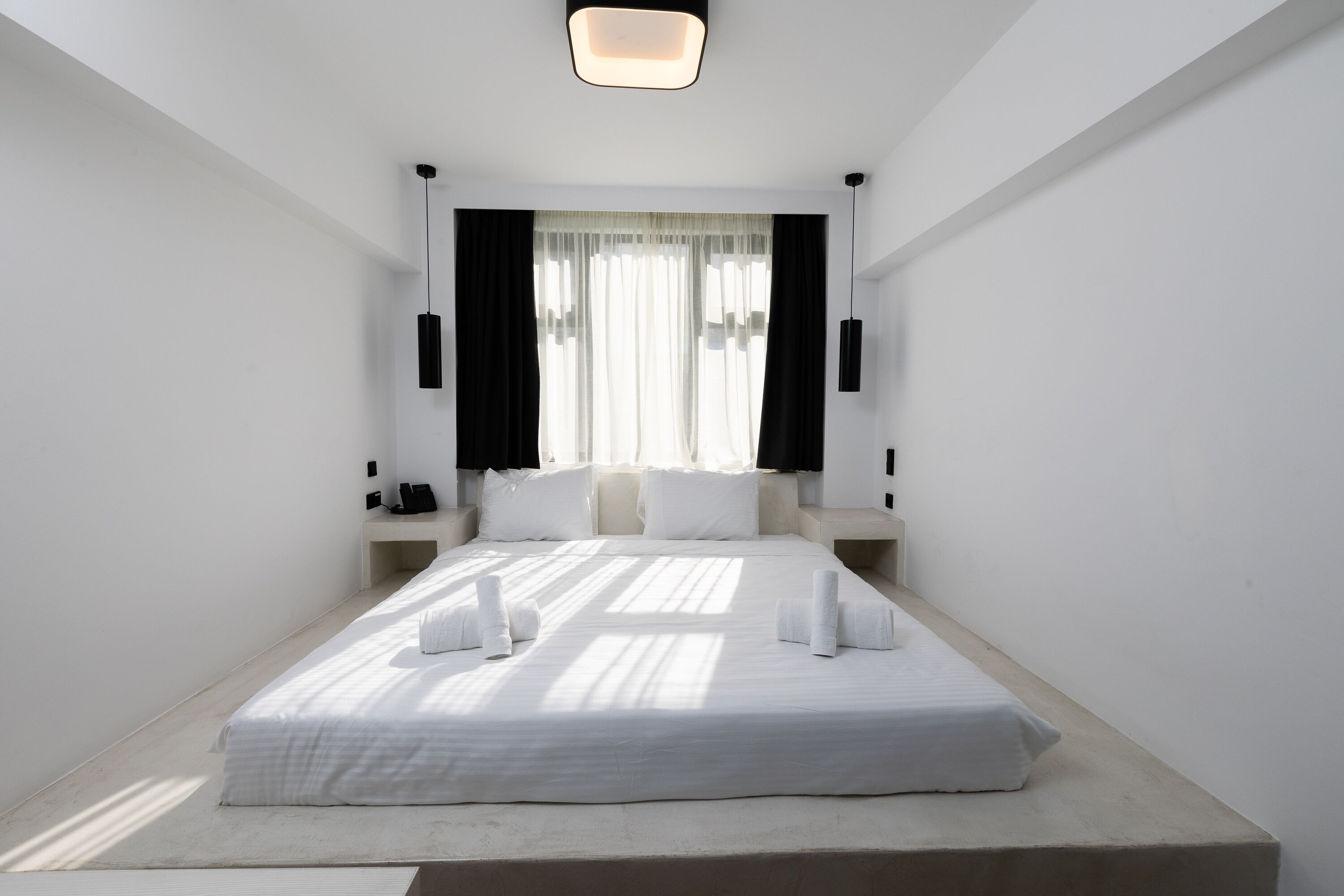 A modern hotel room with a large bed, white linens, a bedside table, a nightstand, a wall-mounted lamp, and sheer curtains.