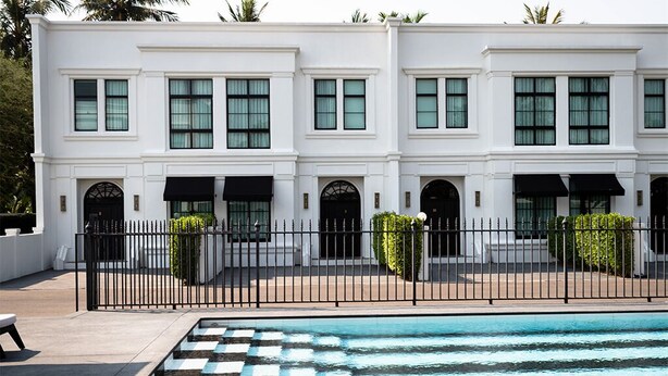 Um edifício branco com várias janelas e toldos pretos, piscina com padrão xadrez e cerca preta.