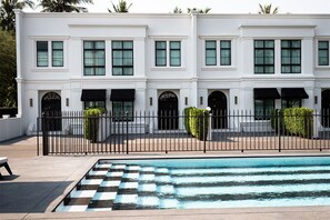 Ein weißes Gebäude mit vielen Fenstern und schwarzen Markisen, ein Pool mit Schachbrettmuster und ein schwarzer Zaun.