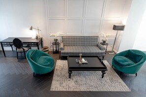 Ein modernes Wohnzimmer mit einem grauen Sofa, zwei türkisfarbenen Sesseln, einem schwarzen Couchtisch, einem kleinen Beistelltisch mit einer Lampe und einem Schreibtisch mit Stuhl.