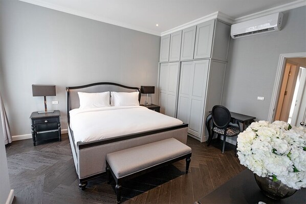 Un dormitorio con cama, mesitas de noche, un banco, una silla, un escritorio y un florero con flores.