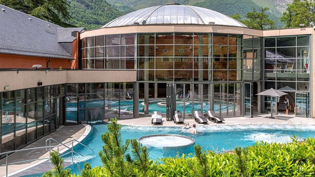 A modern building with a glass facade, a pool area with lounge chairs, and a mountainous backdrop. - Apartment Near Cable Car, Shops & Amenities, East-Facing with Balcony (Cauterets)