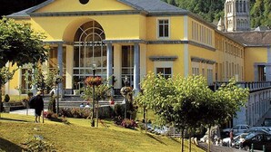 A yellow building with a large arched entrance, a bell tower, and a church in the background. - Apartment Near Cable Car, Shops & Amenities, East-Facing with Balcony (Cauterets)