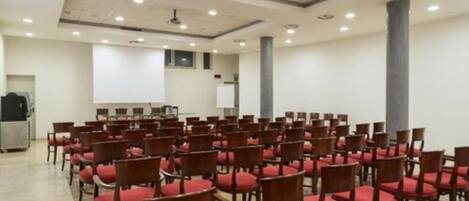 Une salle de conférence avec des rangées de chaises en bois et des coussins rouges, un écran de projection et un tableau blanc.