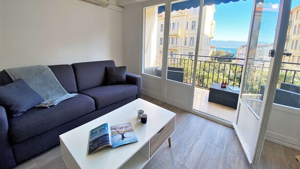 A modern living room with a dark blue sofa, a white coffee table with a magazine, and a large window with a view of buildings and a clear sky. - Ajaccio: Appartement 3 pièces, 6 couchages, 600m de la plage, climatisé, centre-ville (Ajaccio)