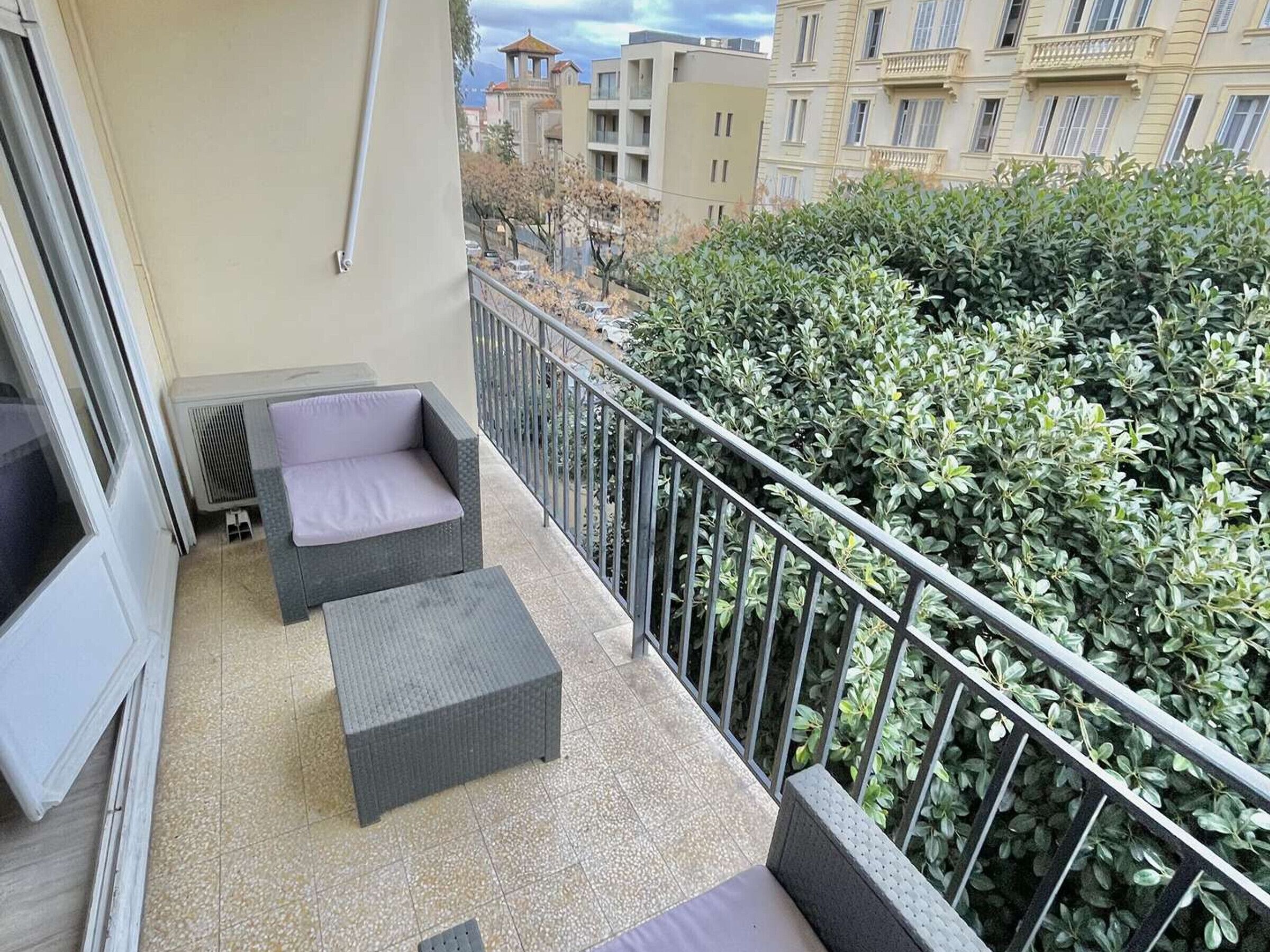 Een balkon met rieten meubilair en een metalen hekwerk, met uitzicht op een begroeide omgeving en gebouwen op de achtergrond.