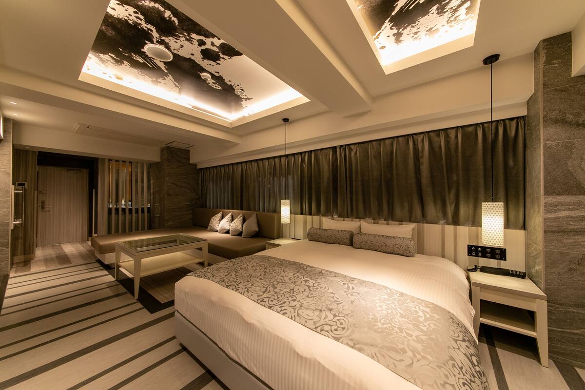 Quarto de hotel moderno com uma cama grande, um sofá, uma mesa de centro de vidro e uma mesa de cabeceira com abajur.