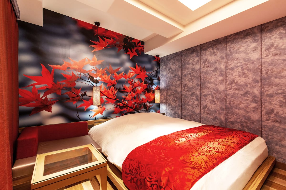 Quarto moderno com uma cama grande, um mural na parede com folhas vermelhas e pretas, e uma mesinha de cabeceira de madeira.