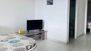 A room with a television on a stand, a small table, and a door leading to a bathroom. - Cozy 1BR Apartment with Parking, 700m from Calvi Beach (Calvi)