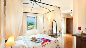 A four-poster bed with a view of a town through a window, a bedside lamp, a mirror, and a wooden dresser with a vase of flowers. - Villa Berry (Corfu)