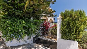 Un edificio blanco con una entrada protegida por una reja, rodeado de abundante vegetación y plantas florecientes.
