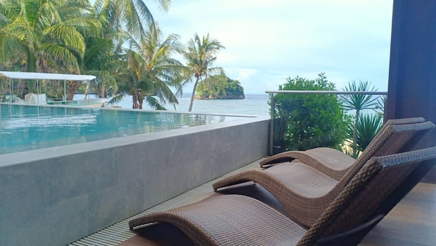 A poolside area with lounge chairs, a view of the ocean, and palm trees.