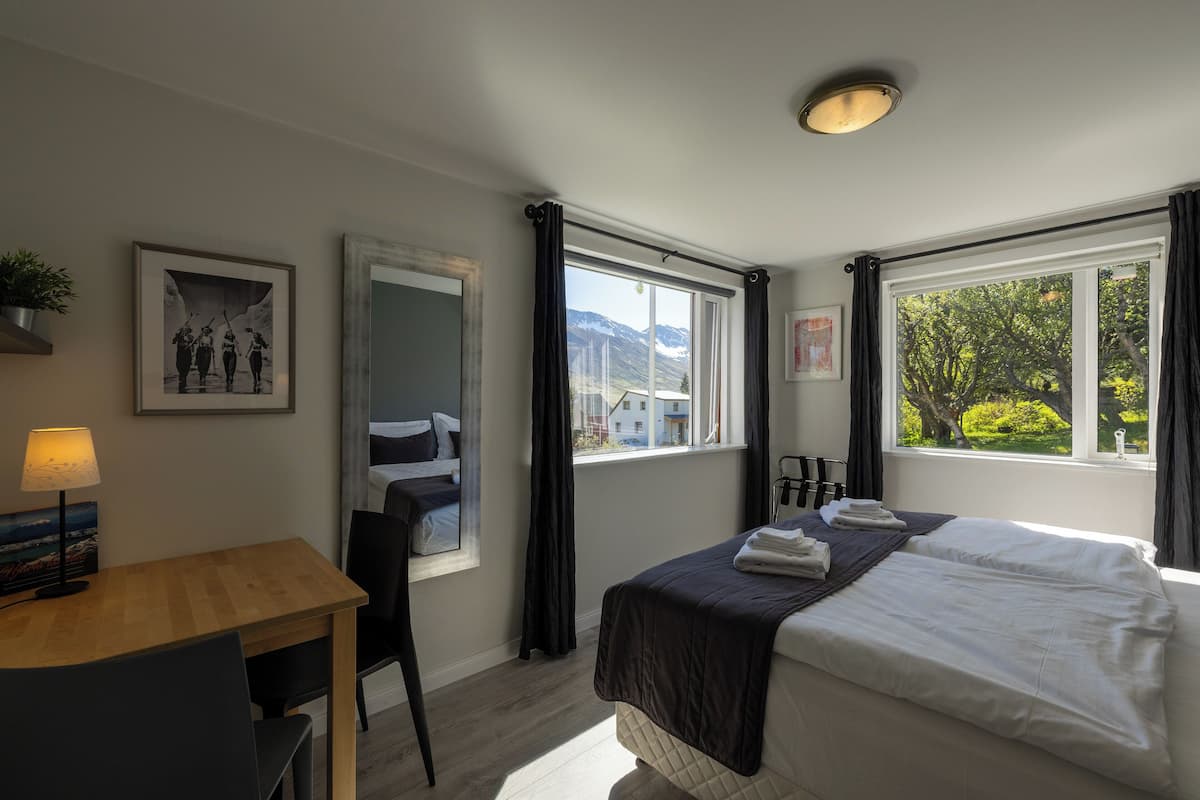 A hotel room with a bed, a desk, a chair, a window with curtains, and a framed picture on the wall.