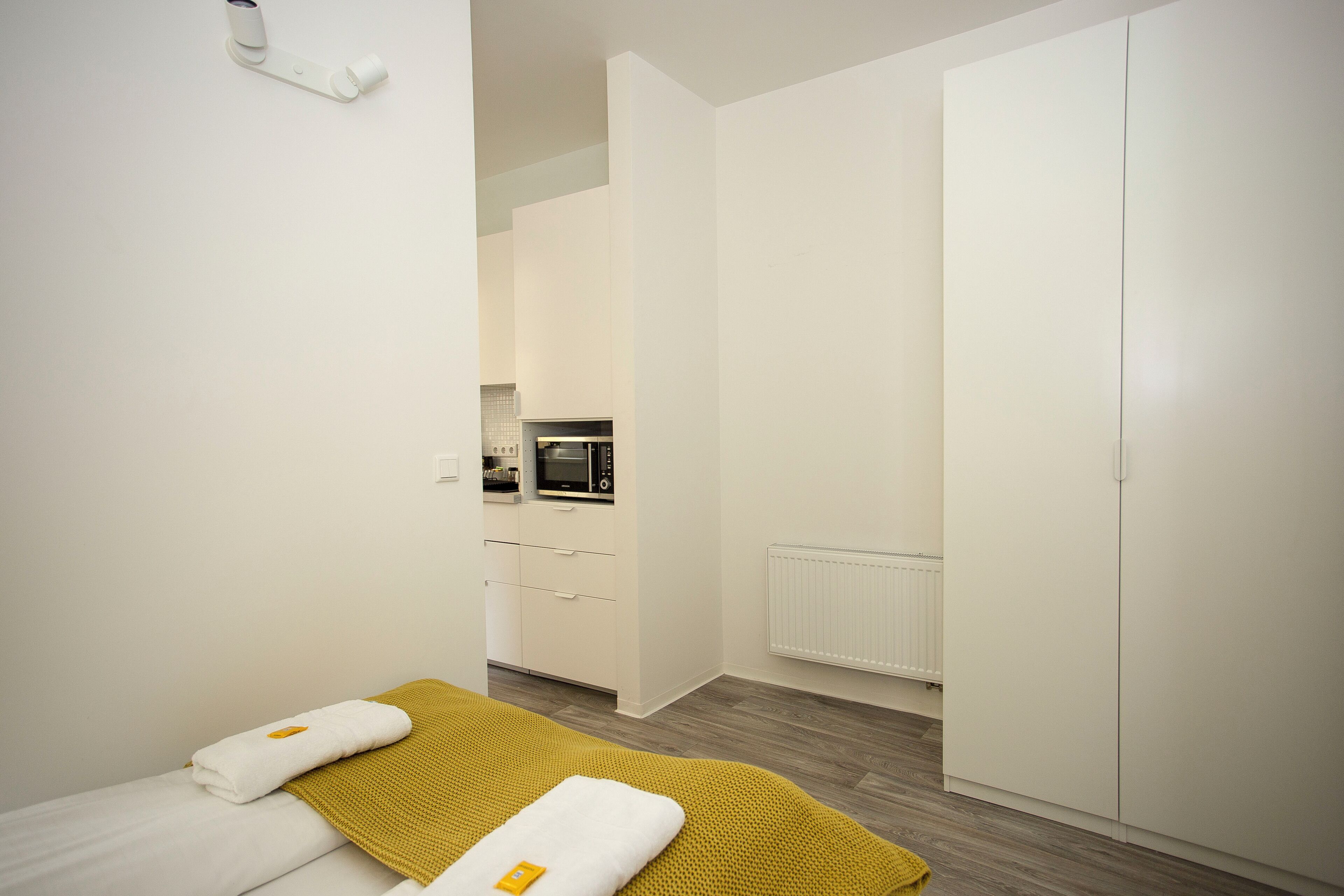 Ein modernes Hotelzimmer mit Bett, gelber Tagesdecke, weißem Kopfkissen, einem kleinen Tisch mit einem gelben Gegenstand und einer Küchenzeile im Hintergrund.