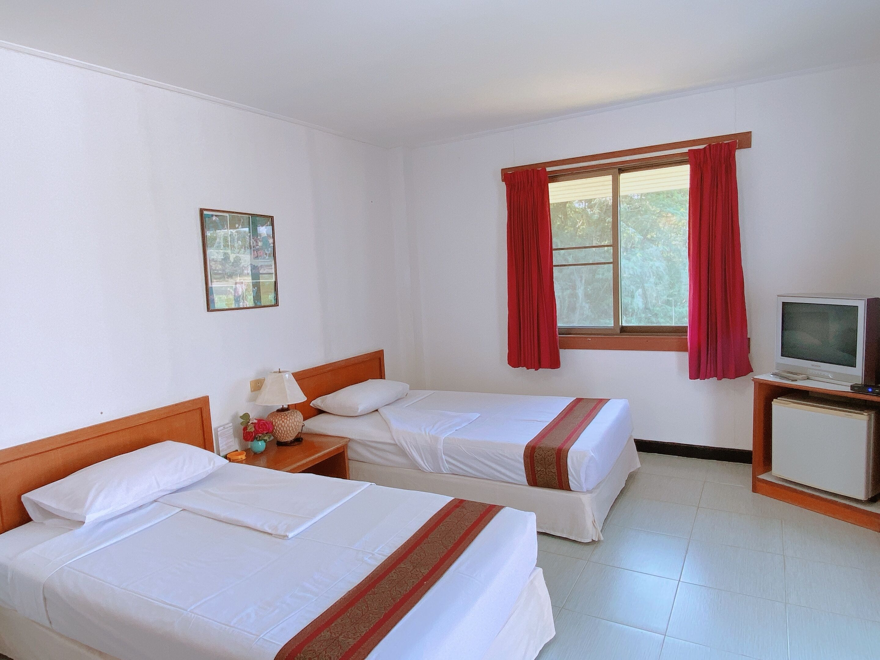 Habitación de hotel con dos camas individuales, televisión, una mesita con lámpara y ventana con cortinas rojas.