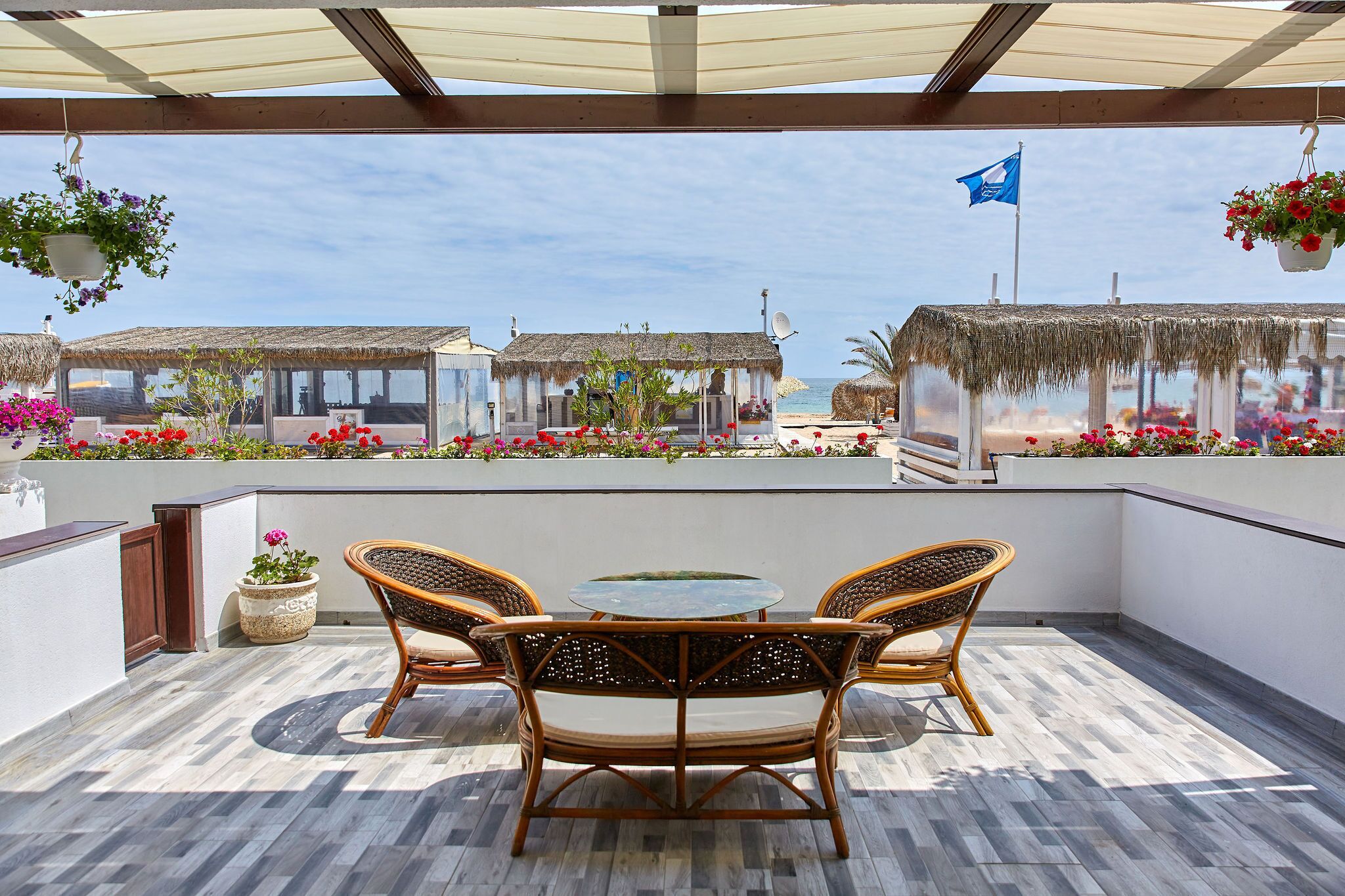 A covered patio with a round table and wicker chairs, overlooking a beach with thatched-roof structures and potted plants.