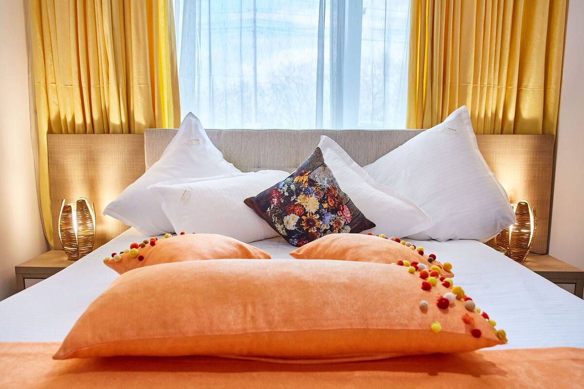Un lit bien fait, agrémenté d’oreillers blancs et orange, d’un coussin à motifs floraux et d’une tête de lit décorative.