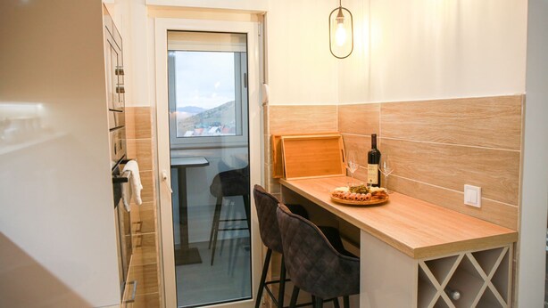 A modern kitchen with a wooden countertop, a wine bottle, and glasses. There is a view through the glass door, and a chair is visible. - Fairy Tale 1 BDR Apartment in City Center (Žabljak)