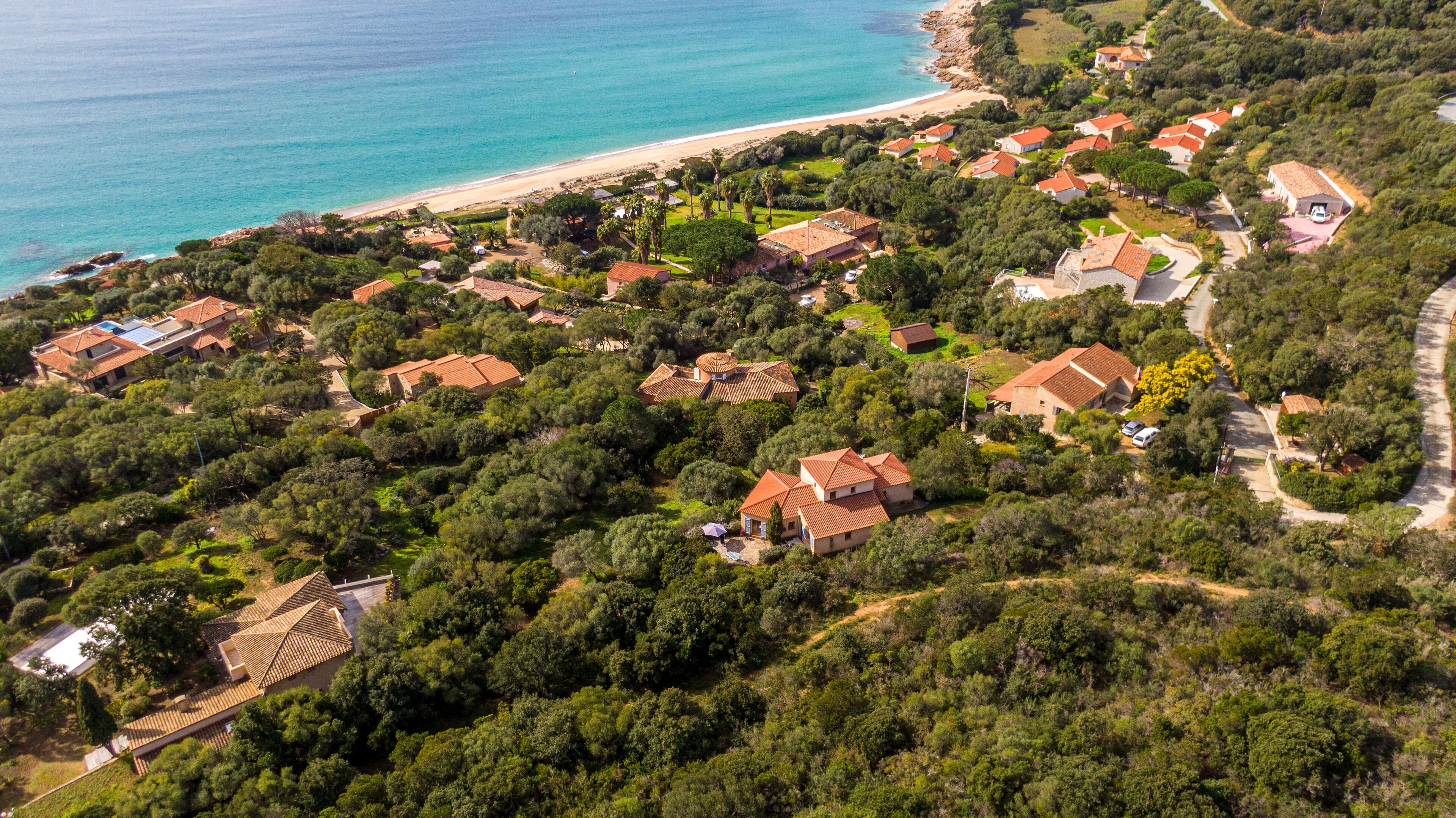 Ett flygbildsbild av ett bostadsområde vid kusten, med hus omgivna av tät vegetation, en strand och en fri utsikt över havet.