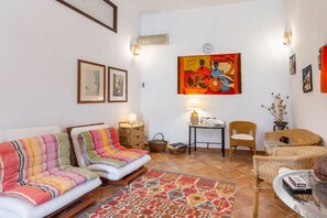 A living room with a sofa, chairs, a patterned rug, a table with a lamp, wicker furniture, and wall art.