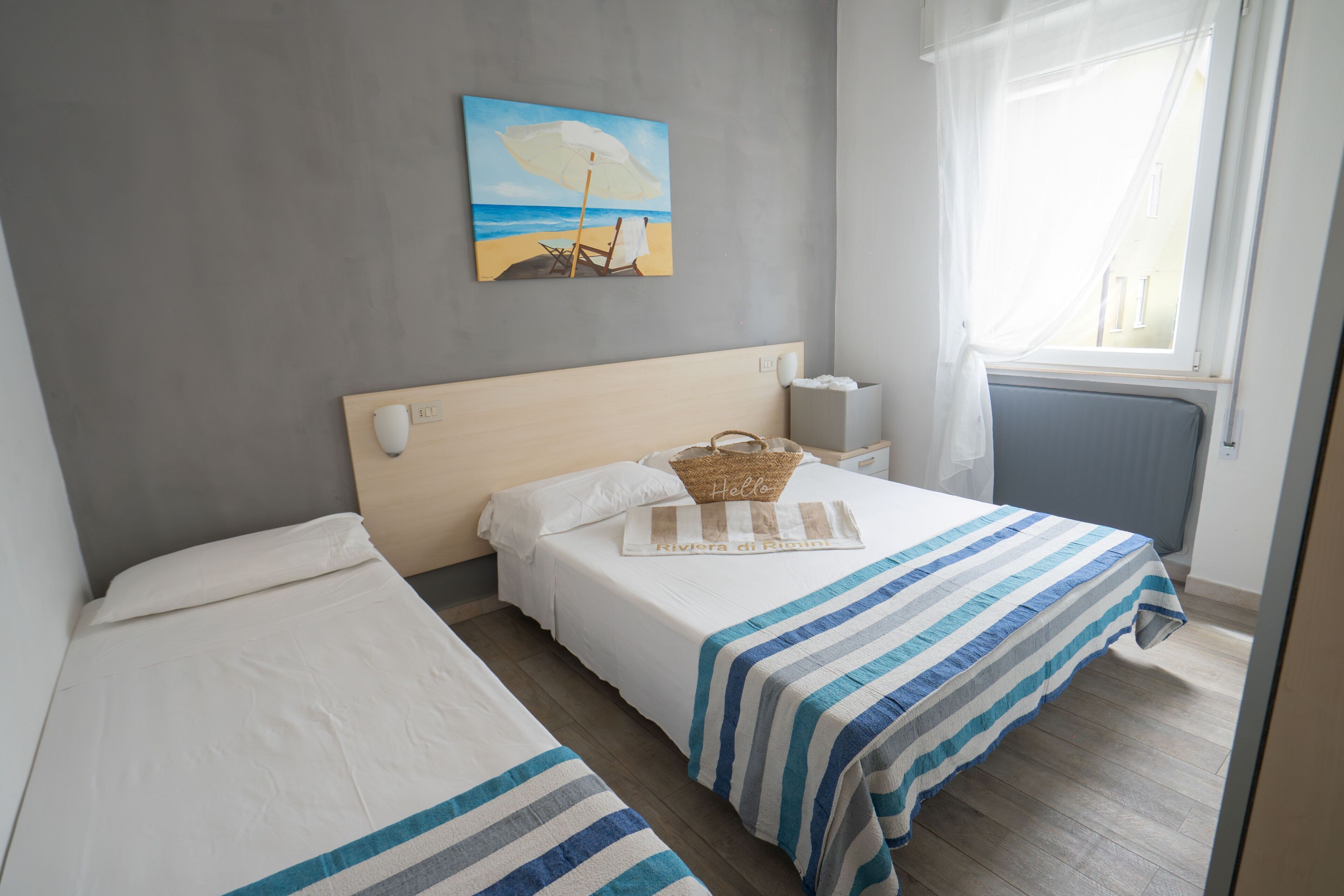 A hotel room with two beds, a beach painting, a window with curtains, and a bedside table with a lamp.