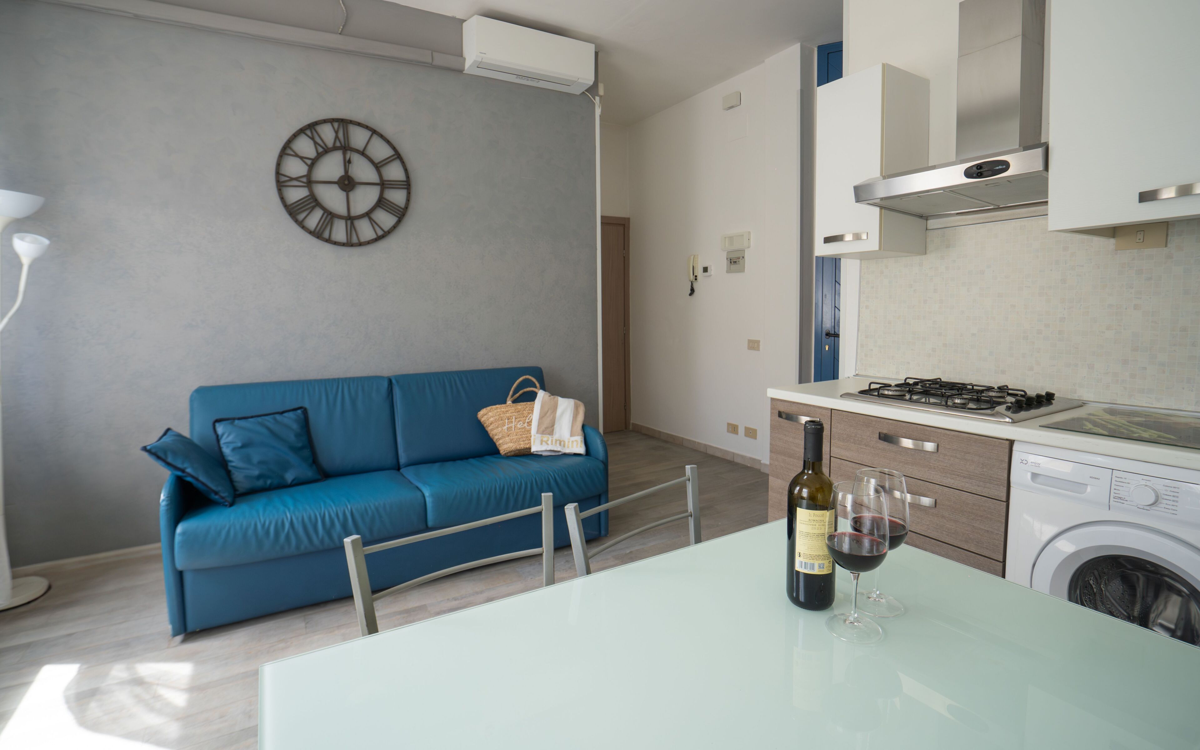 A modern kitchen with a blue sofa, a wall clock, and a bottle of wine on the table.