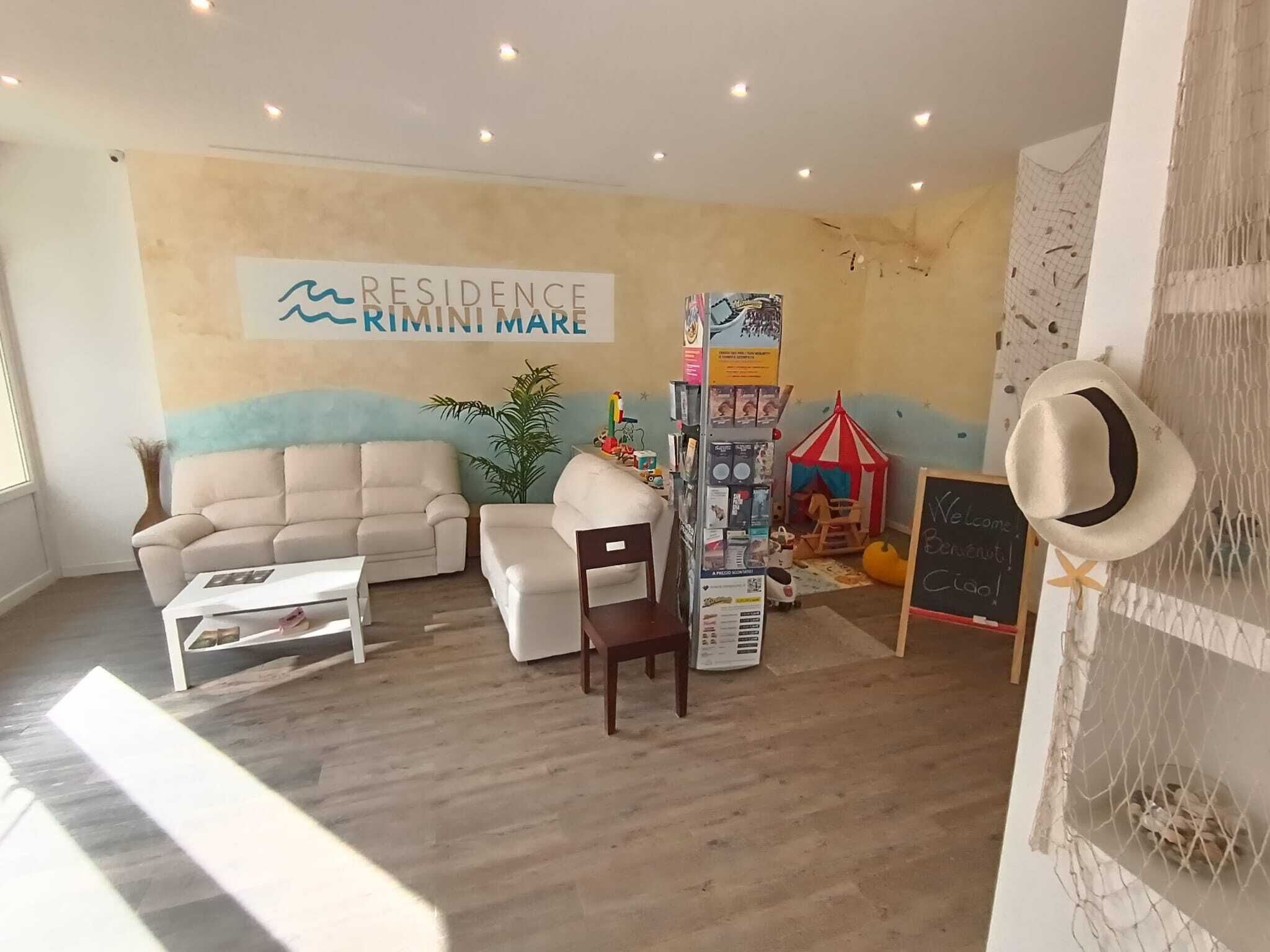 Un salón con sofá, una mesa de centro, una estantería, una mesita con una planta y una pared con un letrero que dice 'RESIDENCE RIMINI MARE'.