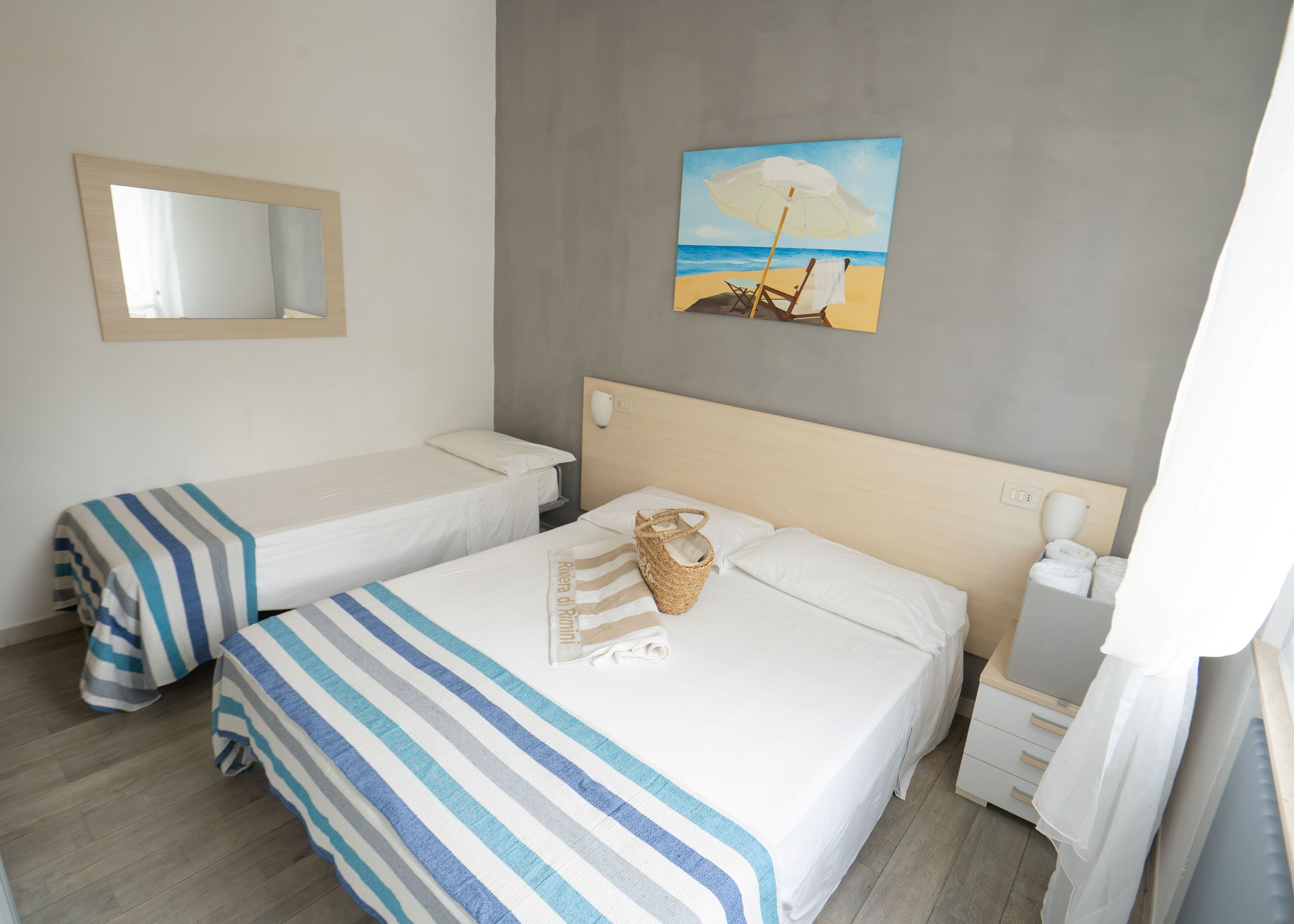 Habitación de hotel con dos camas individuales, un cuadro de una playa, una mesita de noche con una lámpara y un espejo en la pared.