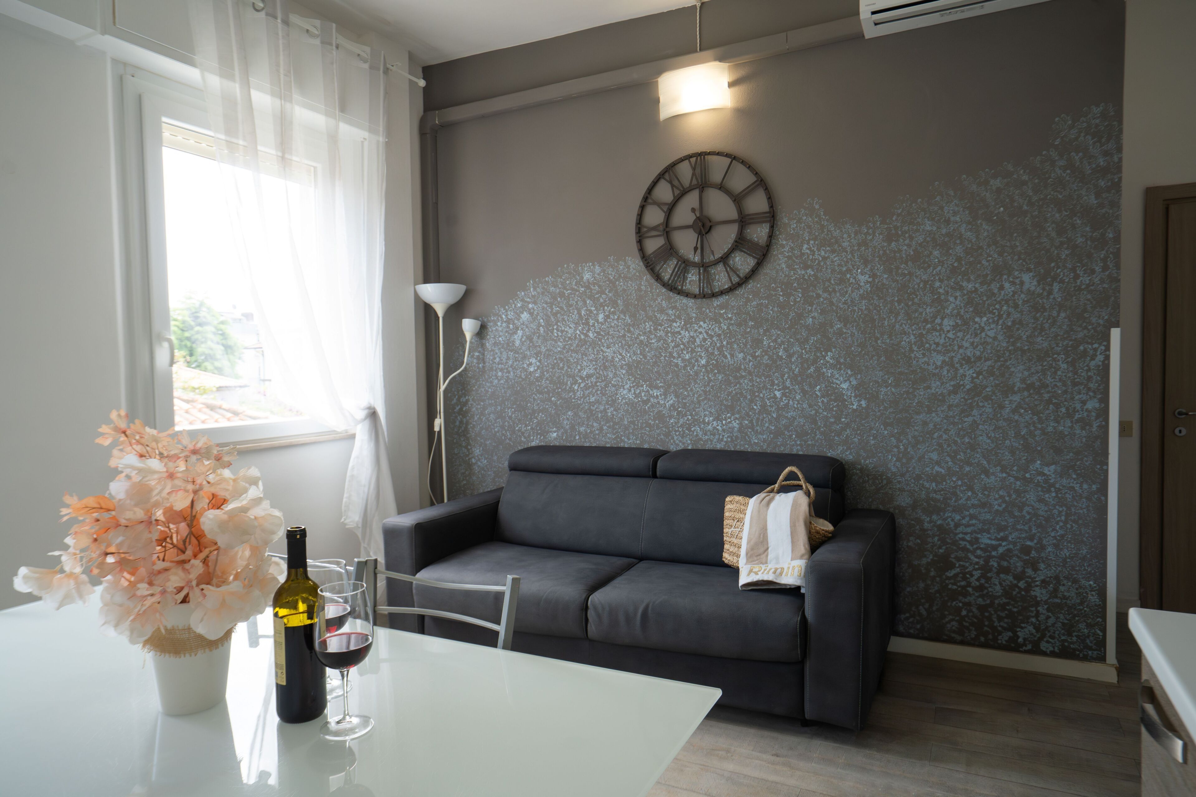 A modern living room with a grey sofa, a wall clock, a vase with flowers, and a bottle of wine on a table.
