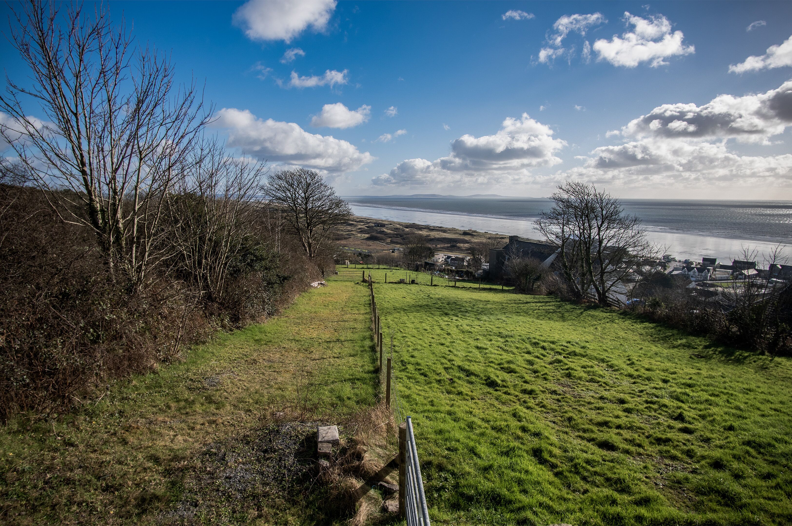 A grassy field with a fence, trees, and a distant view of a body of water and buildings.