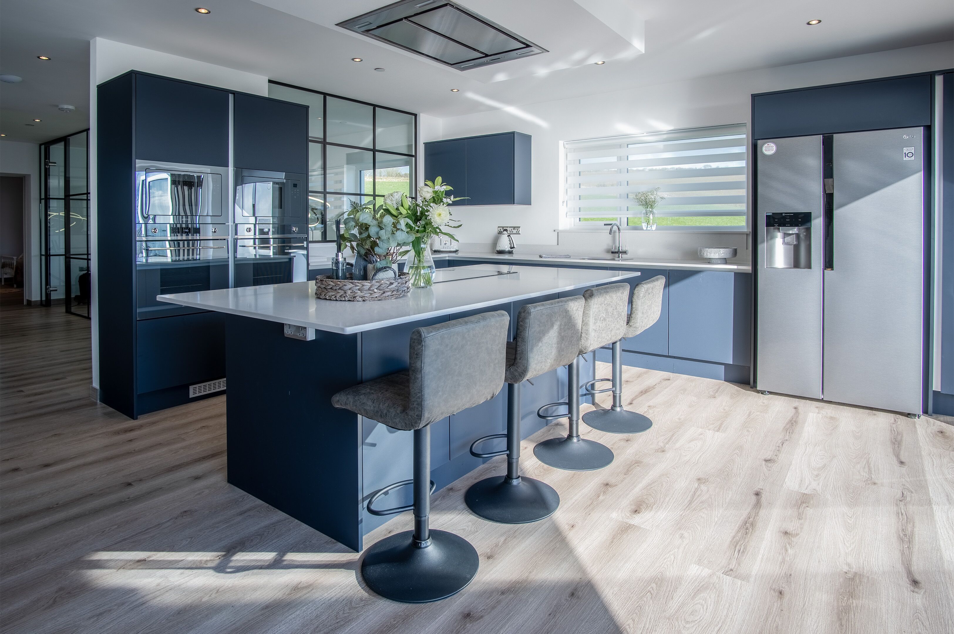 A modern kitchen with a central island, bar stools, a stainless steel refrigerator, and a large window.