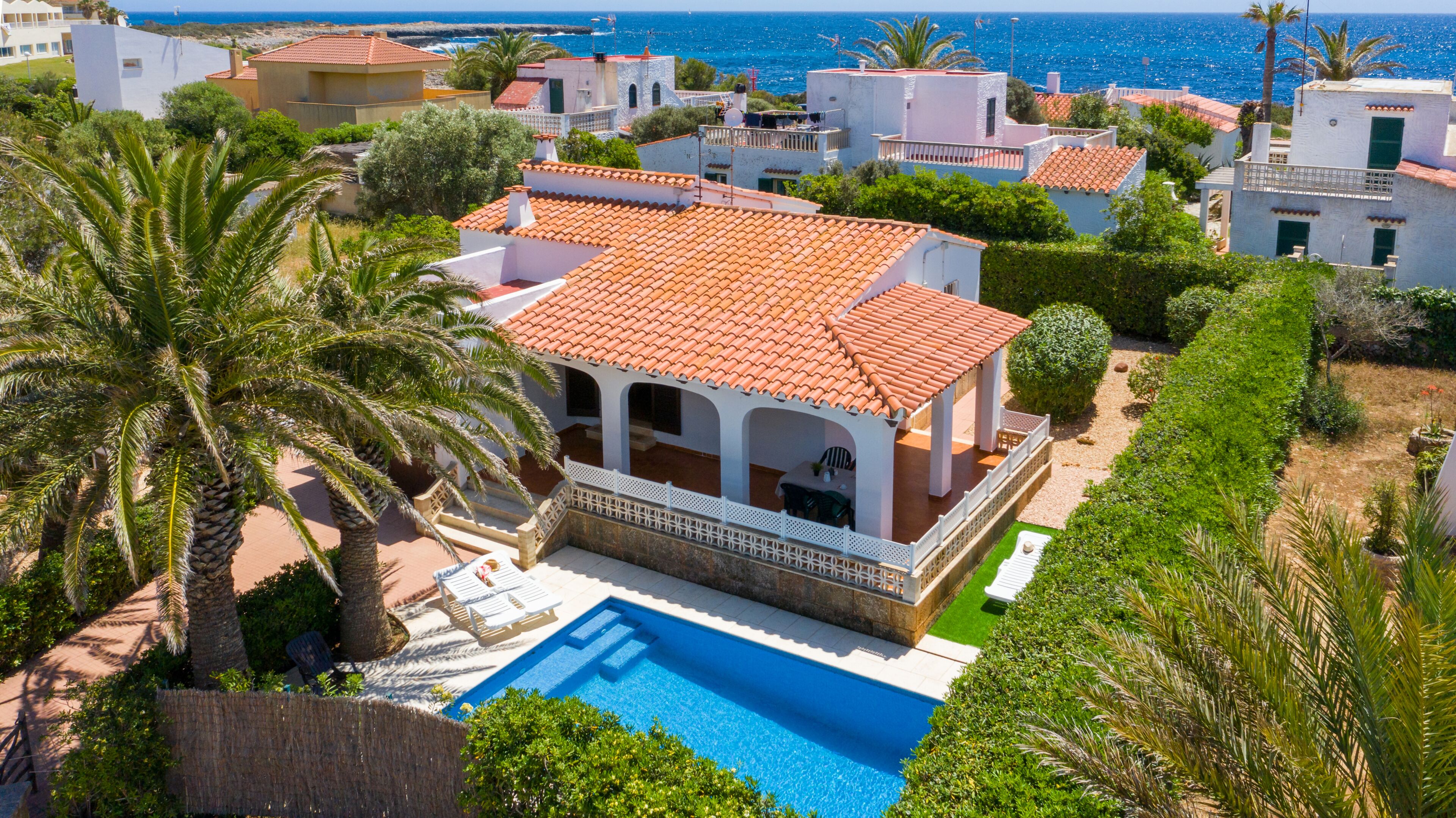 A Mediterranean-style villa with a swimming pool, surrounded by lush greenery and a view of the sea.
