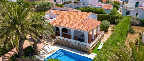 A Mediterranean-style villa with a swimming pool, surrounded by lush greenery and a view of the sea.