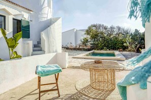 Área externa ensolarada com piscina, mesa de vime e um banquinho de bar.