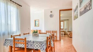 A dining area with a table set for six, wooden chairs, a small plant on the table, and a view into a living room through an open doorway.