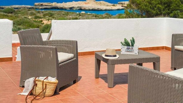 A terrace with wicker furniture, a small table with a hat and a potted plant, and a view of the sea and rocky coastline.