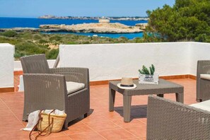 A terrace with wicker furniture, a small table with a hat and a potted plant, and a view of the sea and rocky coastline. - Villa Mascaró | sea views and close to the beach! (Santandria (Platja))