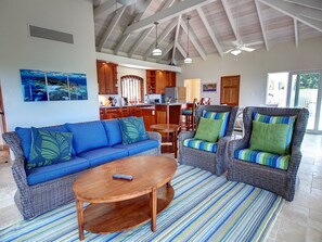 A spacious living area with a blue sofa, two wicker armchairs, a wooden coffee table, and a striped rug. A kitchen is visible in the background with wooden cabinets and a dining area.