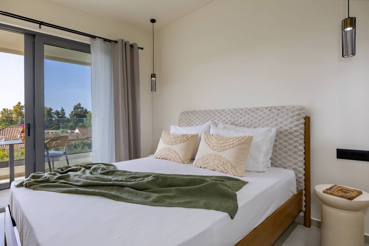 A modern bedroom with a large bed, a view of the outdoors, and a small bedside table.