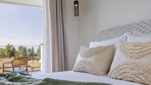 A neatly made bed with white linens and decorative pillows, a green blanket, and a view of a terrace with a chair and table. - Kamara Apartments (Zakynthos)