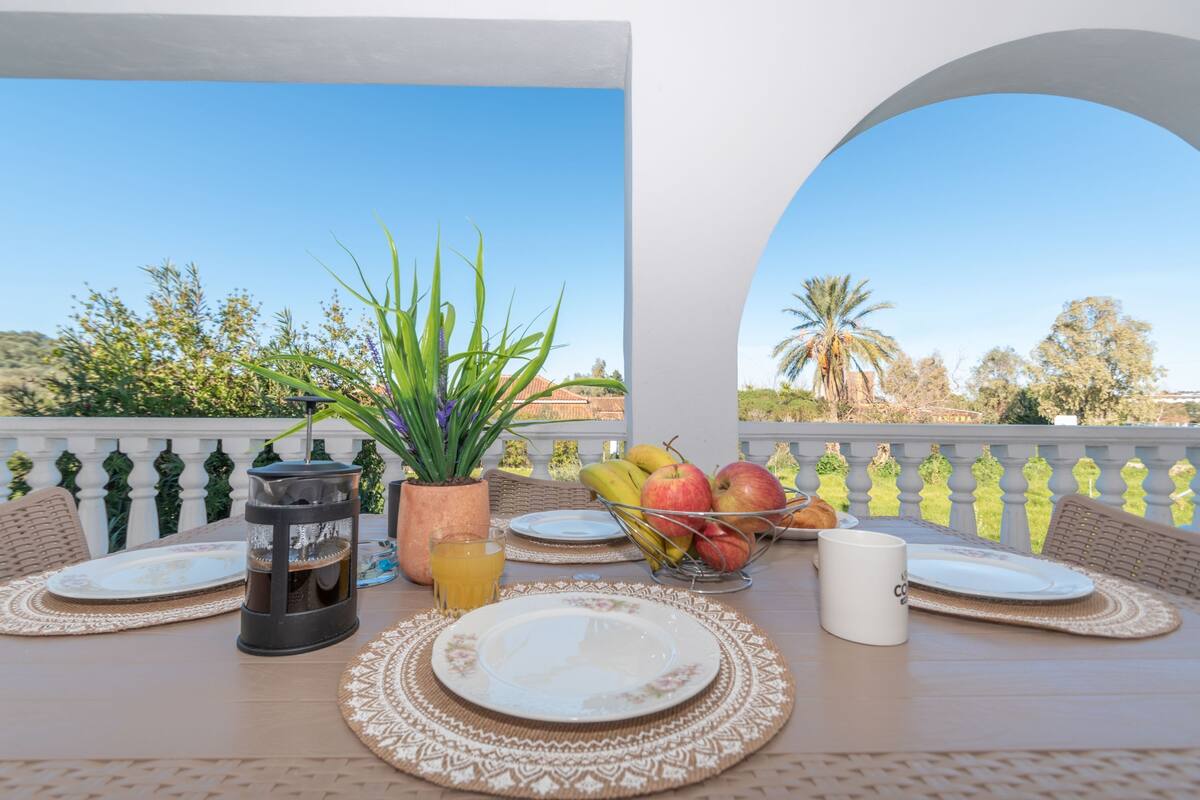 A dining table set with plates, a candle, a fruit bowl, and a glass of orange juice on a balcony with a view of trees and a clear sky.