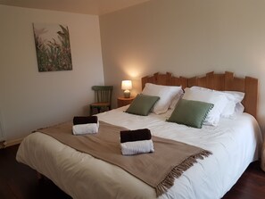 A neatly made bed with white linens and two pillows, a wooden headboard, a small bedside table with a lamp, and a chair.