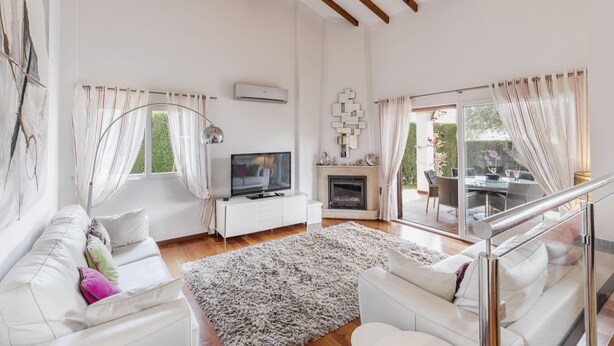 Una sala moderna con un sofá blanco, un televisor de pantalla plana, una chimenea y un comedor visible a través de puertas de vidrio.