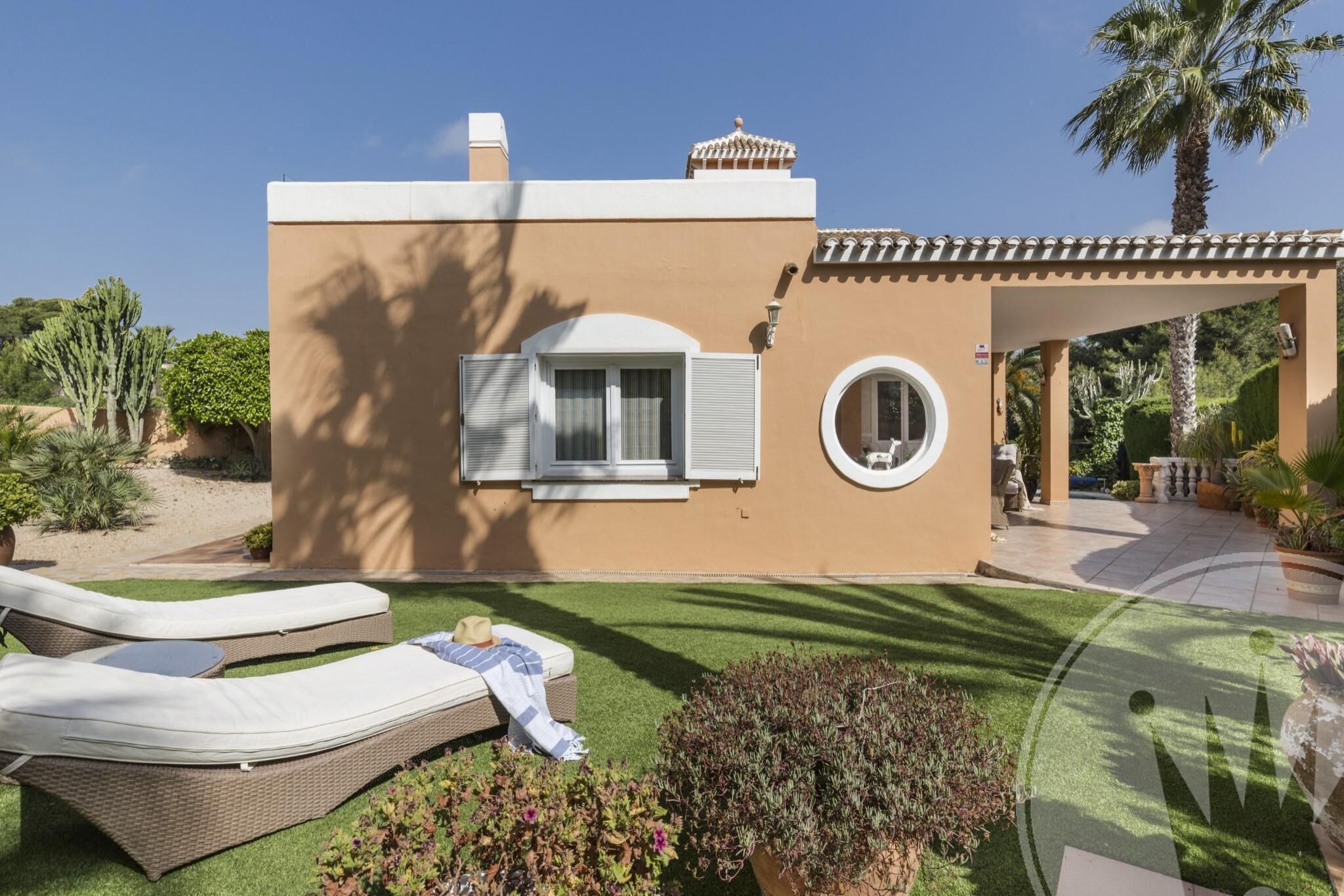 Un bâtiment moderne à la façade beige, doté d’un patio couvert et d’une pelouse bien entretenue avec deux chaises longues.
