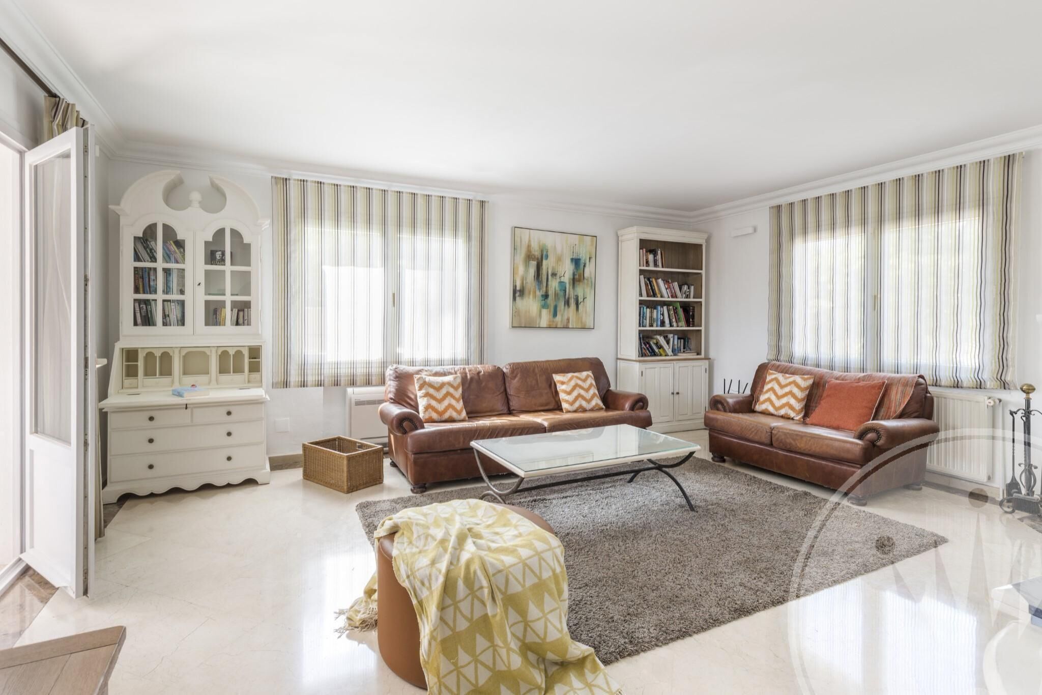 A spacious living room with two brown leather sofas, a glass coffee table, a white cabinet with books, and a striped window treatment.