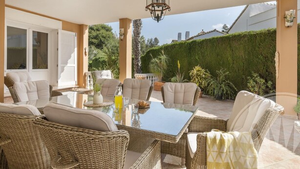 Un patio exterior con mobiliario de mimbre, una mesa de vidrio y un farol.